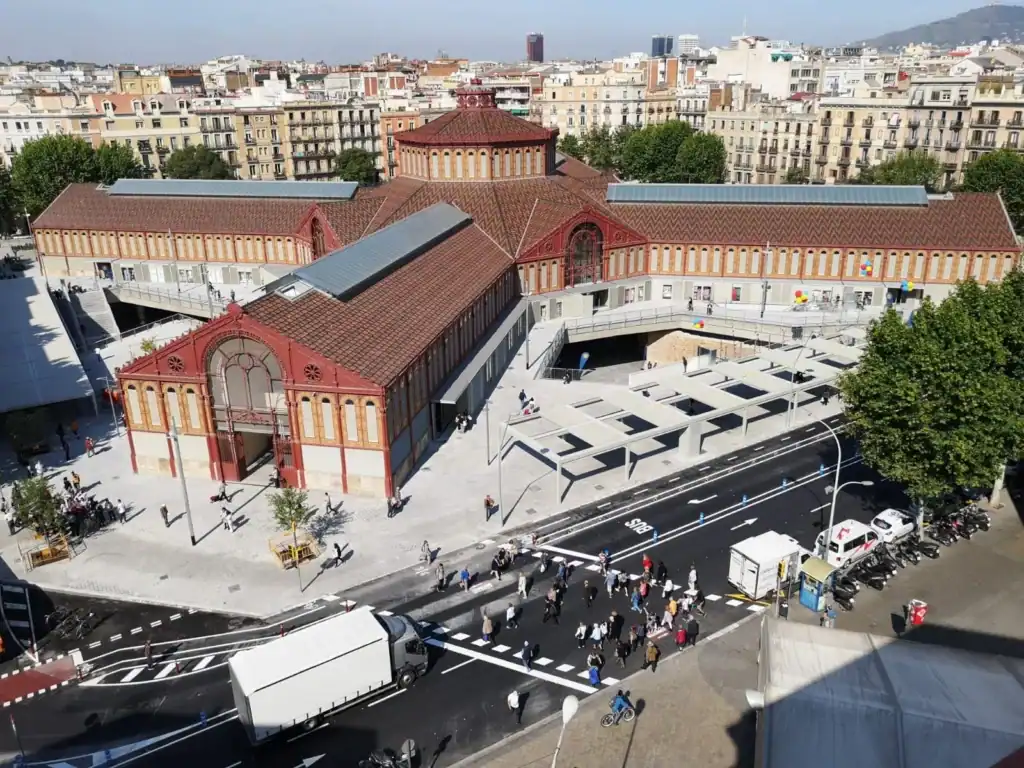 Vista exterior del Mercat Sant Antoni Barcelona, con arquitectura histórica de tejados rojos integrada en un entorno urbano moderno. Se observan peatones, vehículos y señalización vial en una escena que refleja la renovación del barrio.