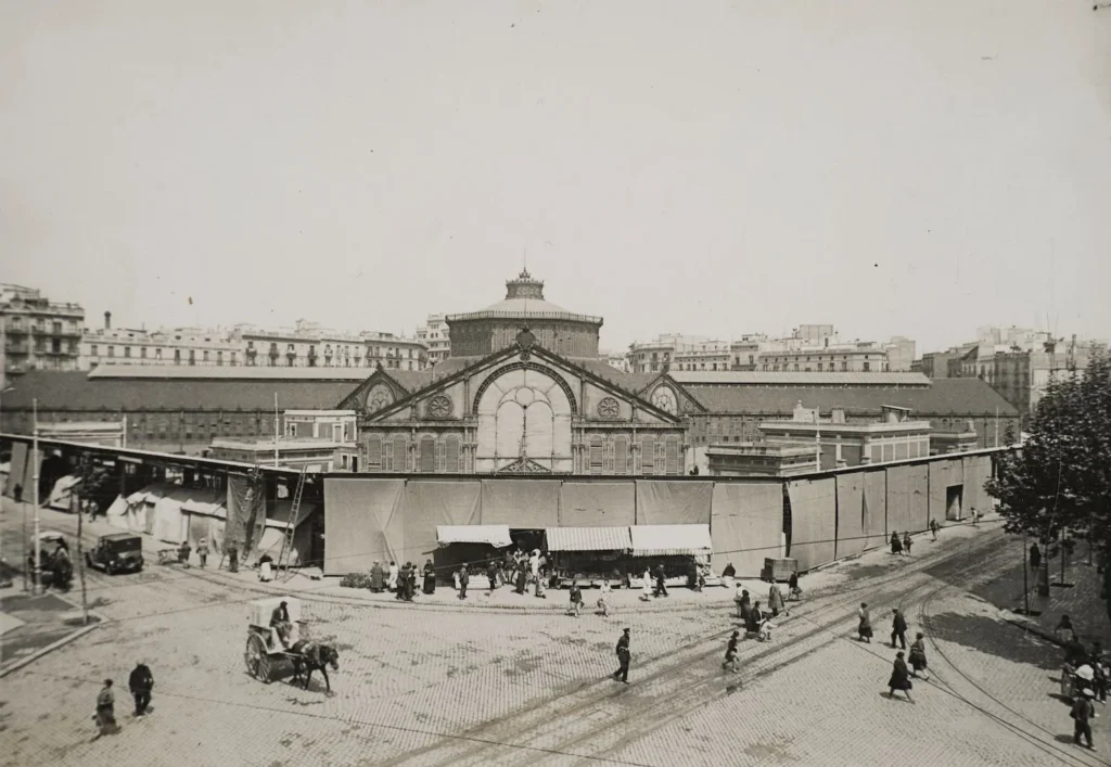 Fotografía histórica del Mercat Sant Antoni Barcelona a principios del siglo XX, con fachada central ornamentada, puestos exteriores y actividad comercial en calles adoquinadas. Se observan peatones, carros de caballos y primeros automóviles en un entorno urbano dinámico.